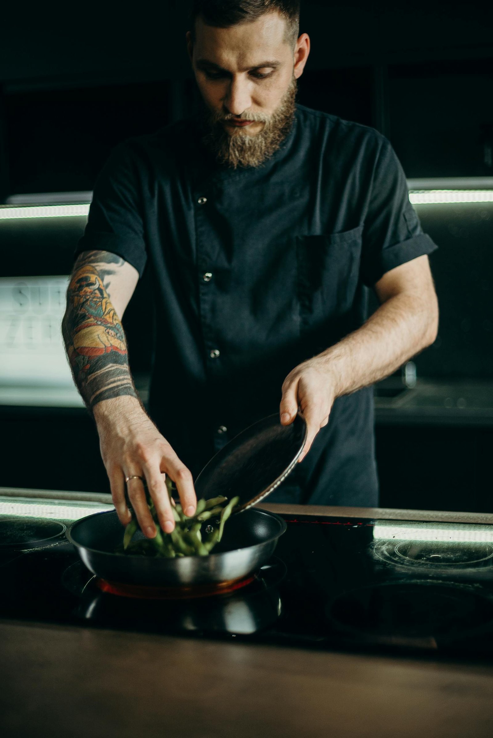 A focused tattooed chef expertly cooks a meal on an indoor stovetop, representing culinary skill.